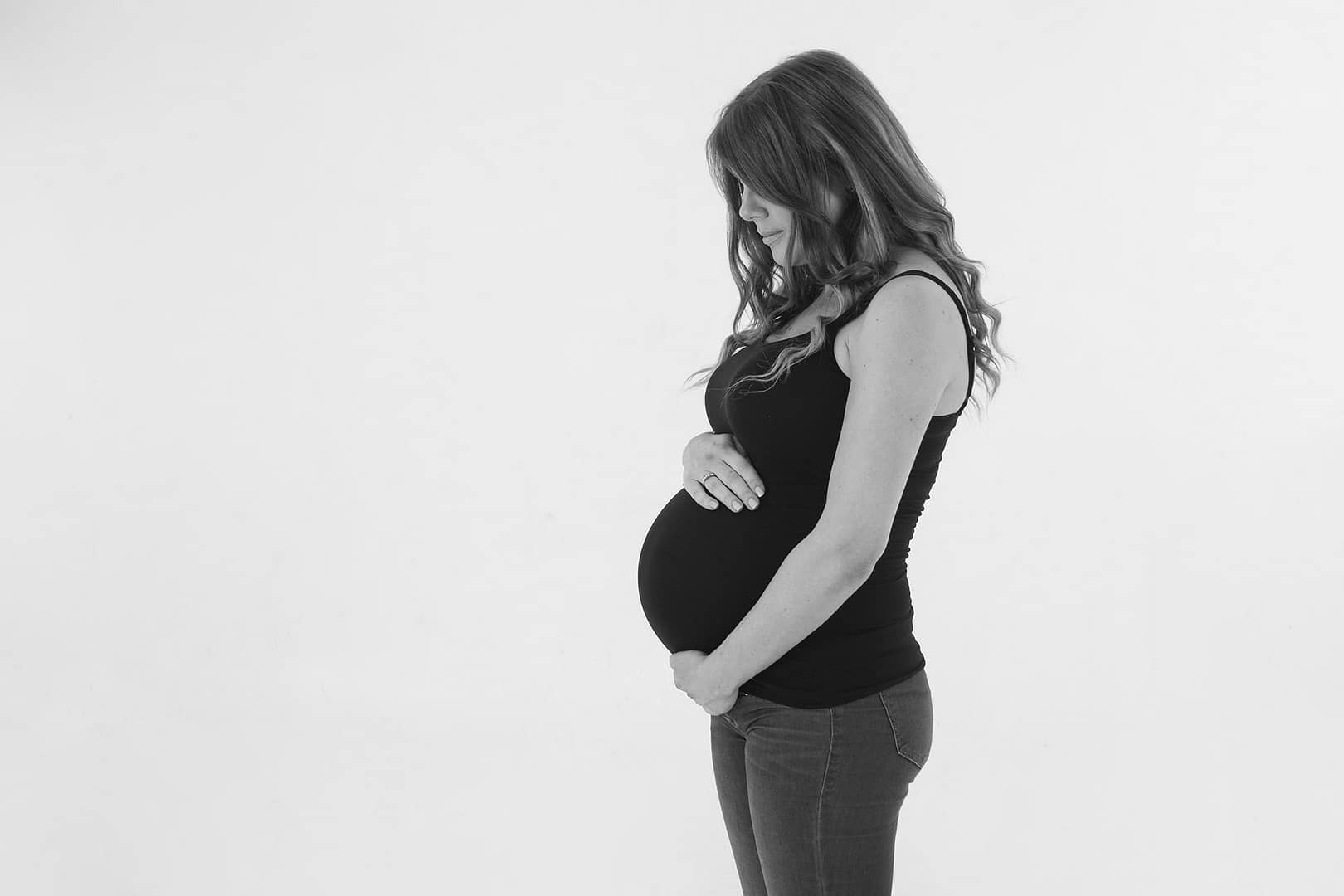 Pregnant mamma-to-be standing infront of a white wall.  She's turned to the side, facing the left of the image.  She's looking down and cradling her bump
