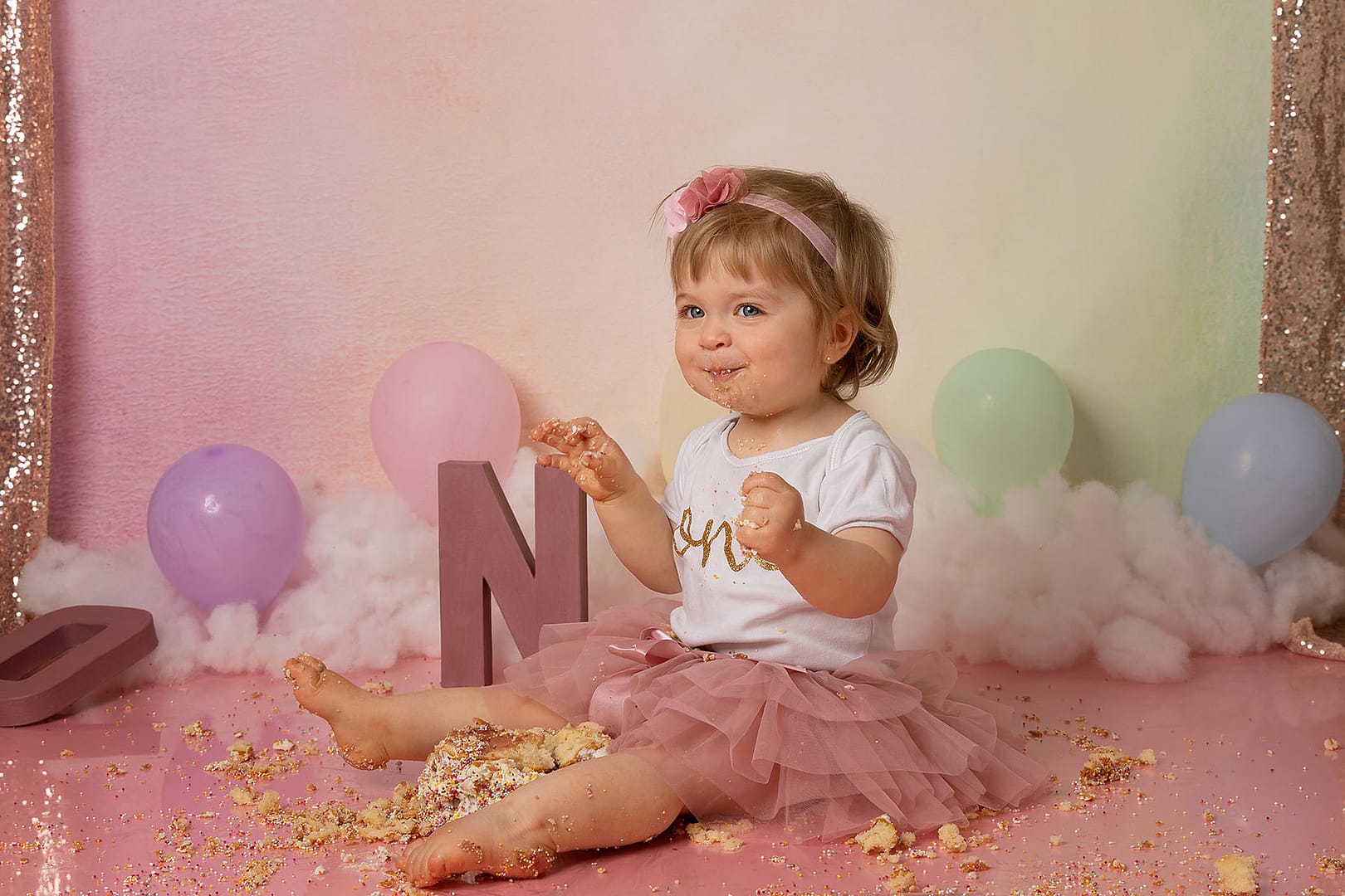 rainbow cake smash theme with little one year old girl surrounded by lots of cake pieces as she enjoys more of her cake.  By cake smash photographer Sutton Coldfield birmingham