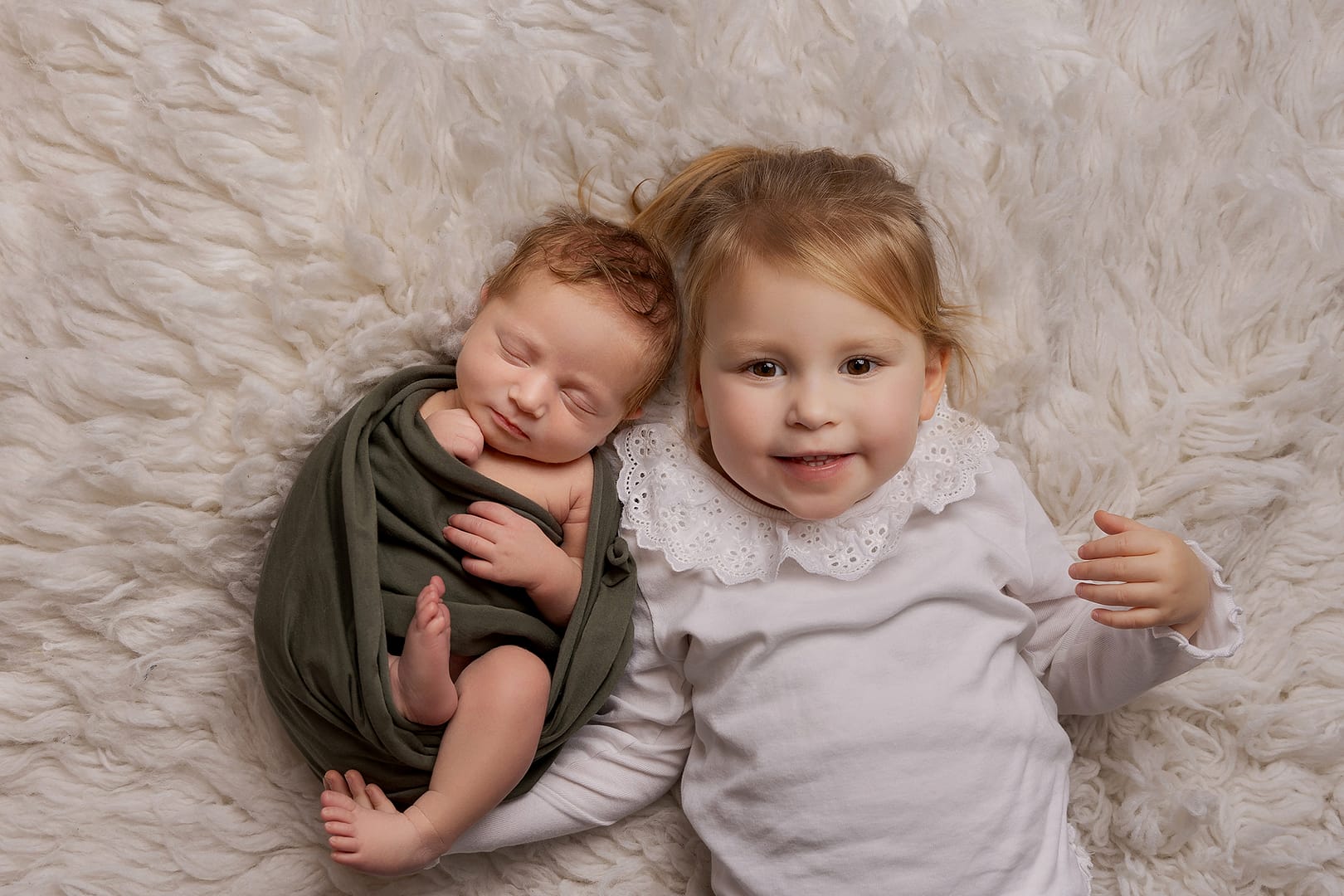 Newborn baby boy with his sister posing on a cream rug. Baby is wrapped in a dark khaki green wrap and big sister is wearing a white top and smiling proudly up at the camera. Newborn photograph taken by Nicki Byrne Photography, newborn photographer birmingham
