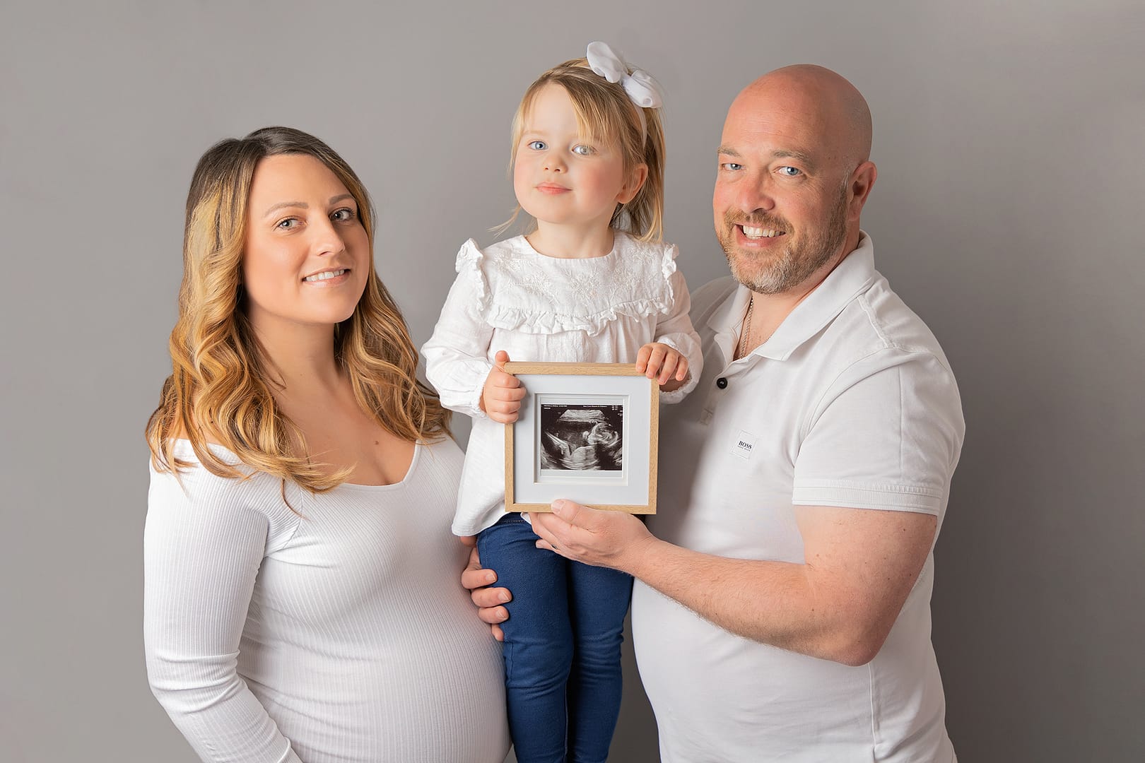 Pregnant mother-to-be with her eldest daughter and husband all wearing white tops and denim jeans.  Little girl is holding her brother's scan photo.  Taken by award winning maternity photographer, Nicki Byrne Photography, maternity photographer birmingham, maternity photographer Sutton Coldfield 