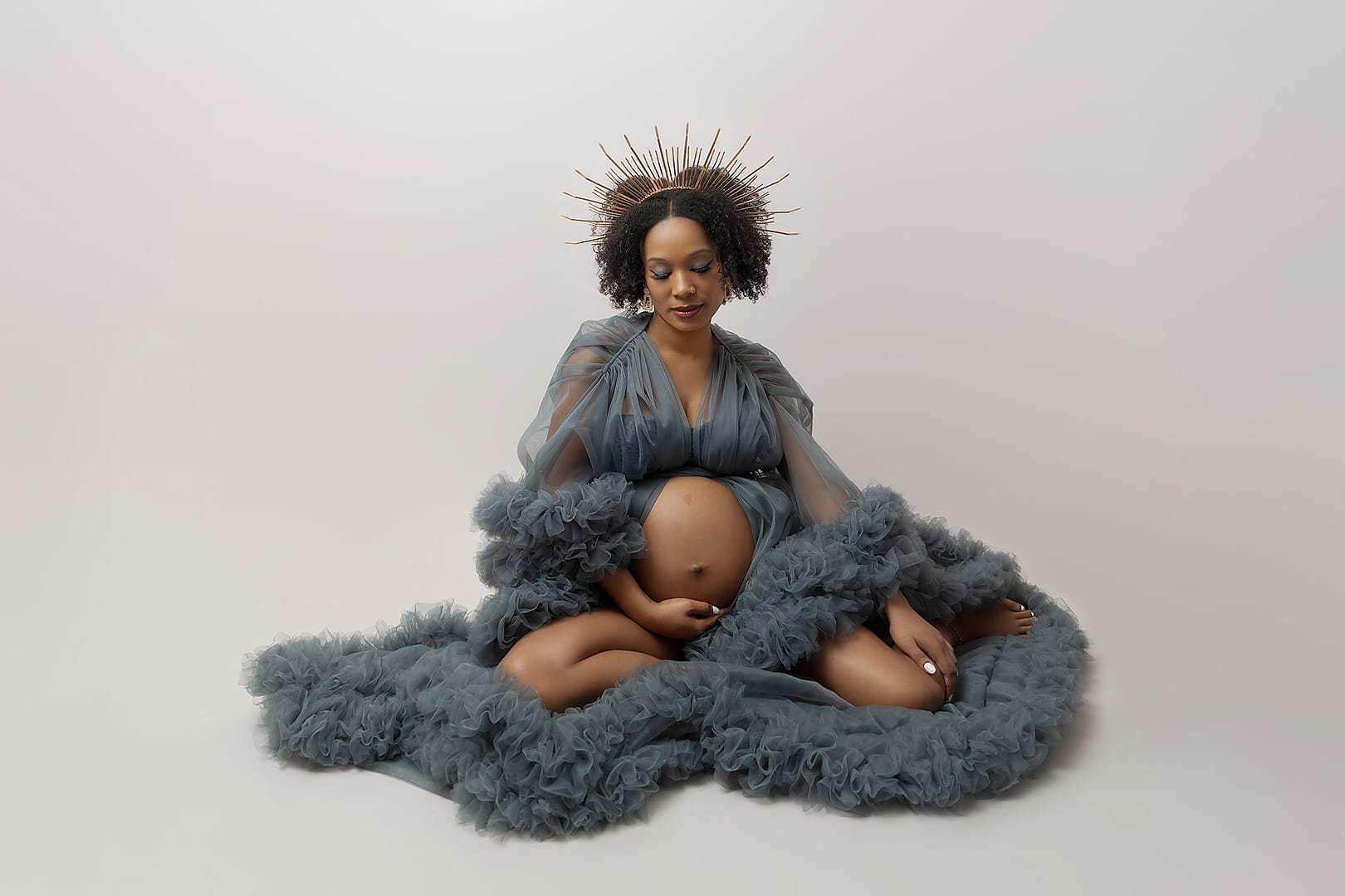 Beautiful pregnant queen showcasing her beautiful pregnancy bump wearing a petrol blue tulle maternity gown.  Mamma-to-be is wearing a gold crown.  She is sat on the floor delicately holding her bump.  Taken by award winning maternity photographer Nicki Byrne Photography, maternity photographer Sutton Coldfield birmingham