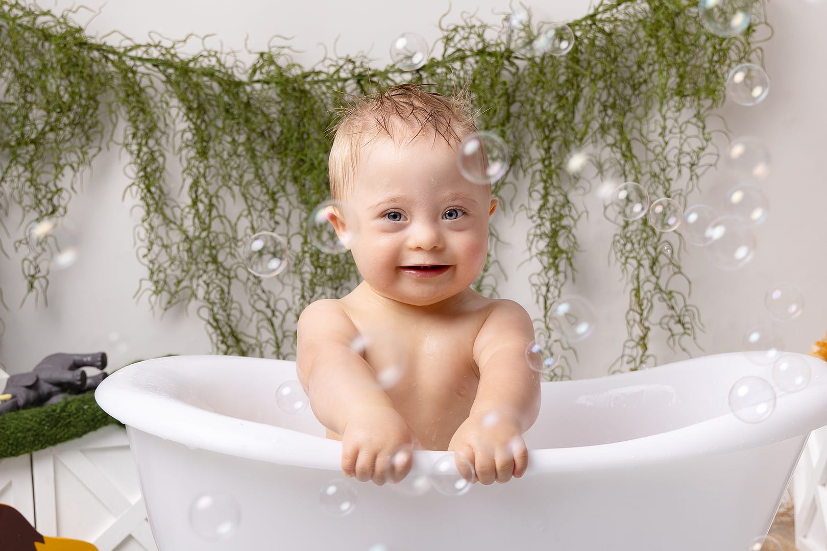 Gorgeous boy smiling at the camera whilst in a roll top bath surrounded by bubbles.  Taken during his cake smash with cake smash photographer Nicki Byrne Photography, Sutton Coldfield Birmingham