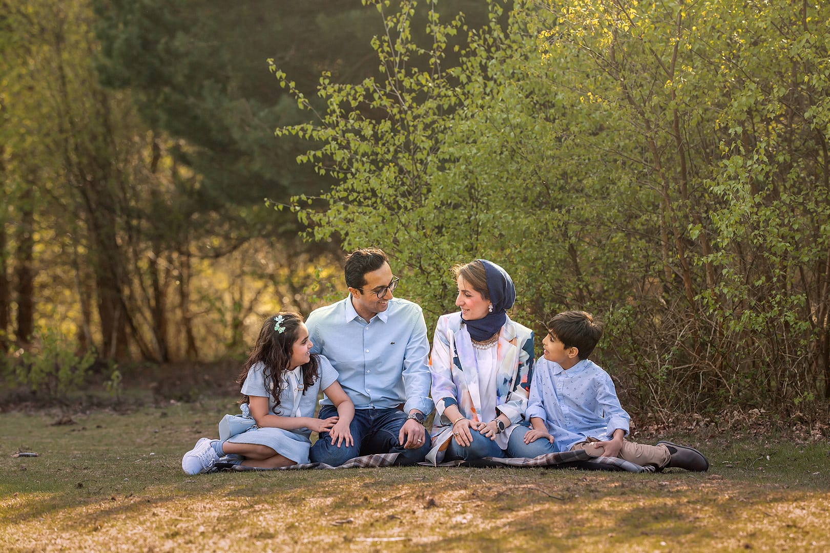 Outdoor family photoshoot set in the summer, warm sun rays are peaking through the trees as a family of four dressed in shades of blue are sat naturally talking together.  Taken by Nicki Byrne Photography, family photographer Sutton Coldfield Birmingham