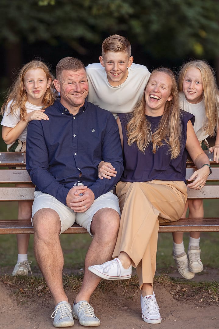 Family of 5 laughing naturally sat around a park bench.  Taken by outdoor family photographer Nicki Byrne Photography, Sutton Coldfield Birmingham