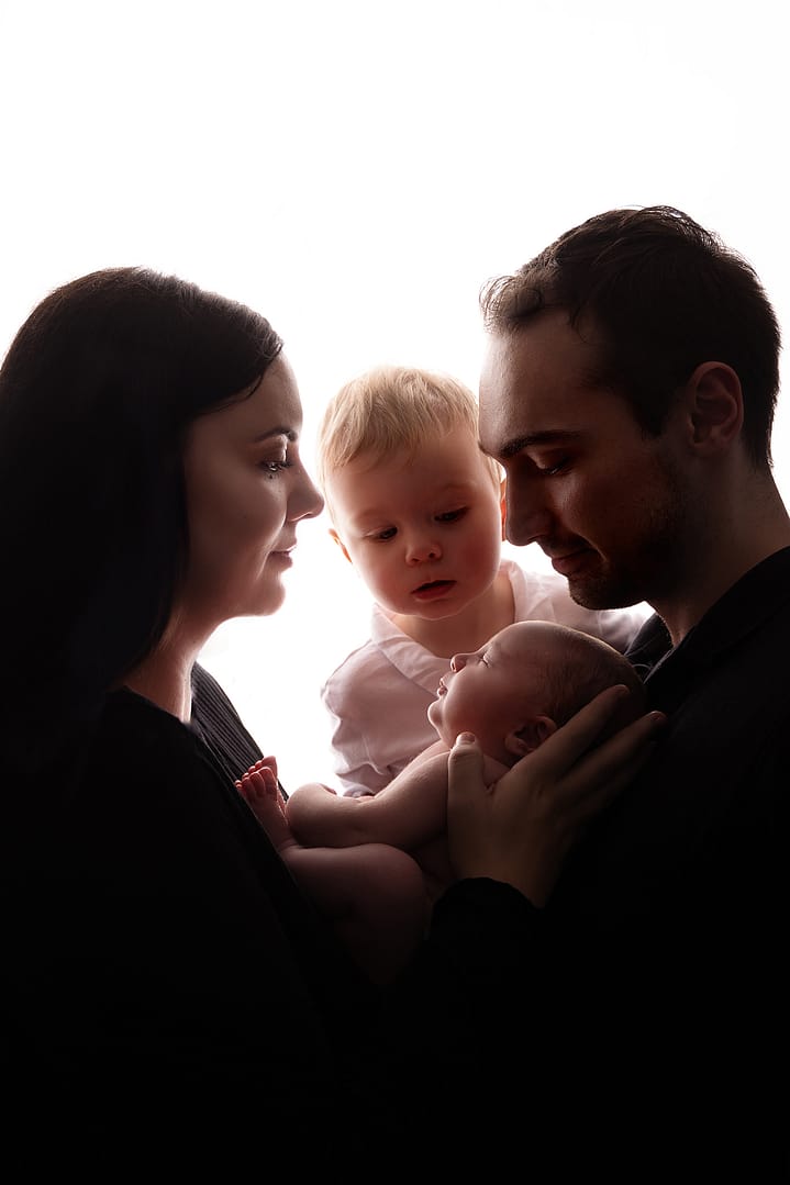 A back lit image where mom and dad are holding their newborn and toddler brother is looking over dad's shoulder at his baby sister. Taken by newborn photographer Nicki Byrne Photography, newborn photographer birmingham
