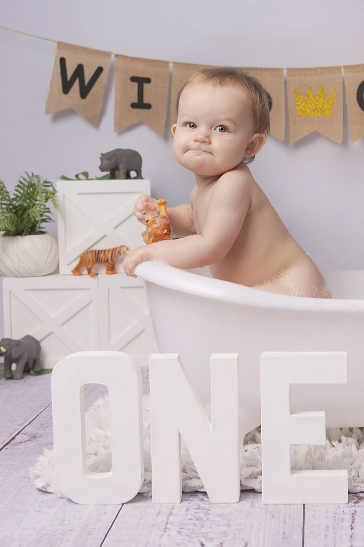 One year old celebrating with a post cake smash bubble bath, she is climbing out of the side of the bath.  Big O N E letters are at the bottom of the image to signify her first birthday.  Taken at Nicki Byrne Photography, cake smash photographer Sutton Coldfield, birmingham