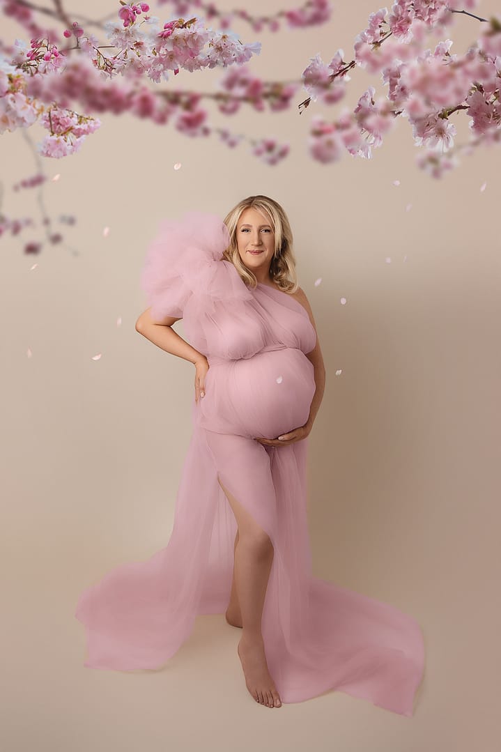 Maternity photo - mama-to-be dressed in a soft pink tulle dress with one hand on her hip.  Background is a light cream colour and there is pink blossom at the top of the image.  Taken by award winning maternity photographer, Nicki Byrne Photography, maternity photographer Sutton Coldfield birmingham