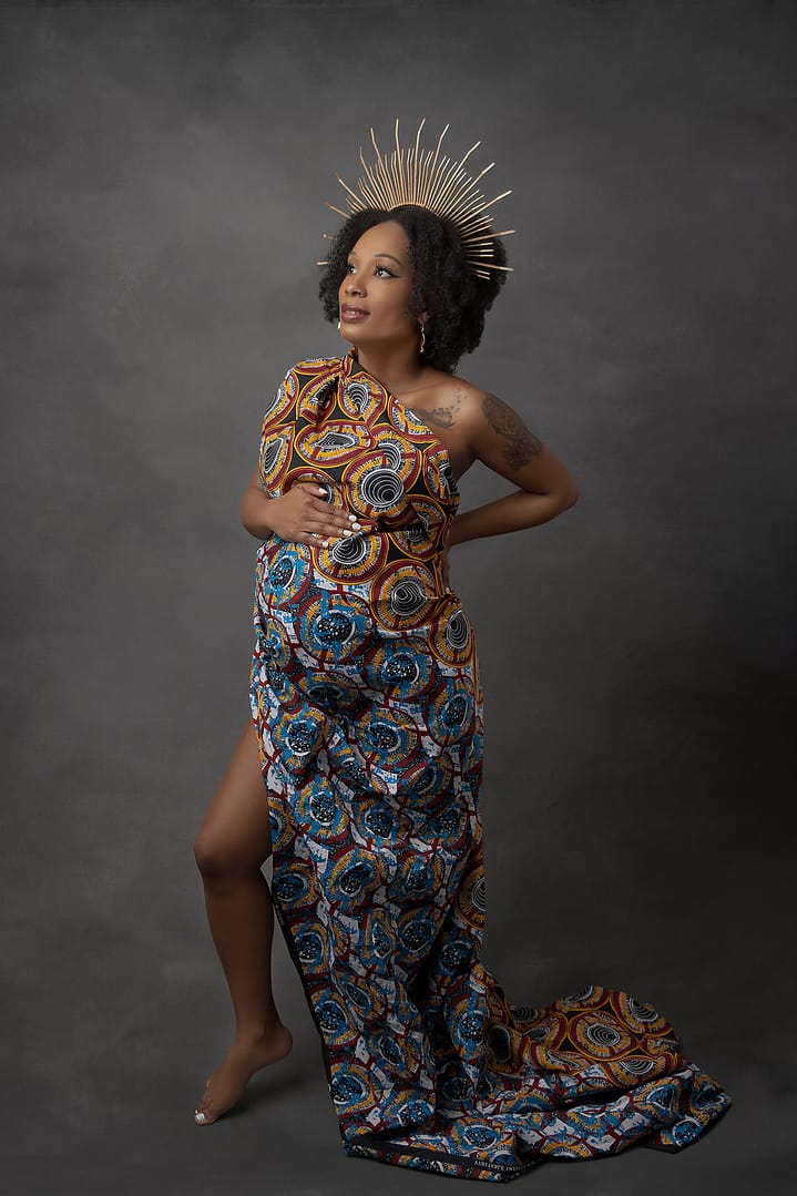 Beautiful pregnant mother-to-be wearing a traditional African material gown and a golden crown.  She is holding her bump with one hand and her hip with the other.  Taken by award winning maternity photographer Nicki Byrne Photography, maternity photographer Sutton Coldfield birmingham