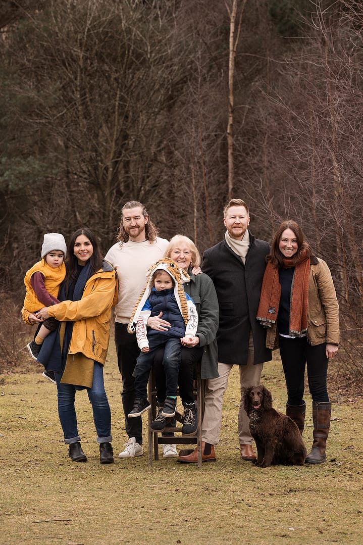 Large extended family posing for Nicki Byrne Photography at Sutton park.  It is winter and everyone is dressed in coats, wellies and a scarf/hat.  They have a dog in their photo too.  Taken by Nicki Byrne Photography, Family photographer Sutton Coldfield Birmingham