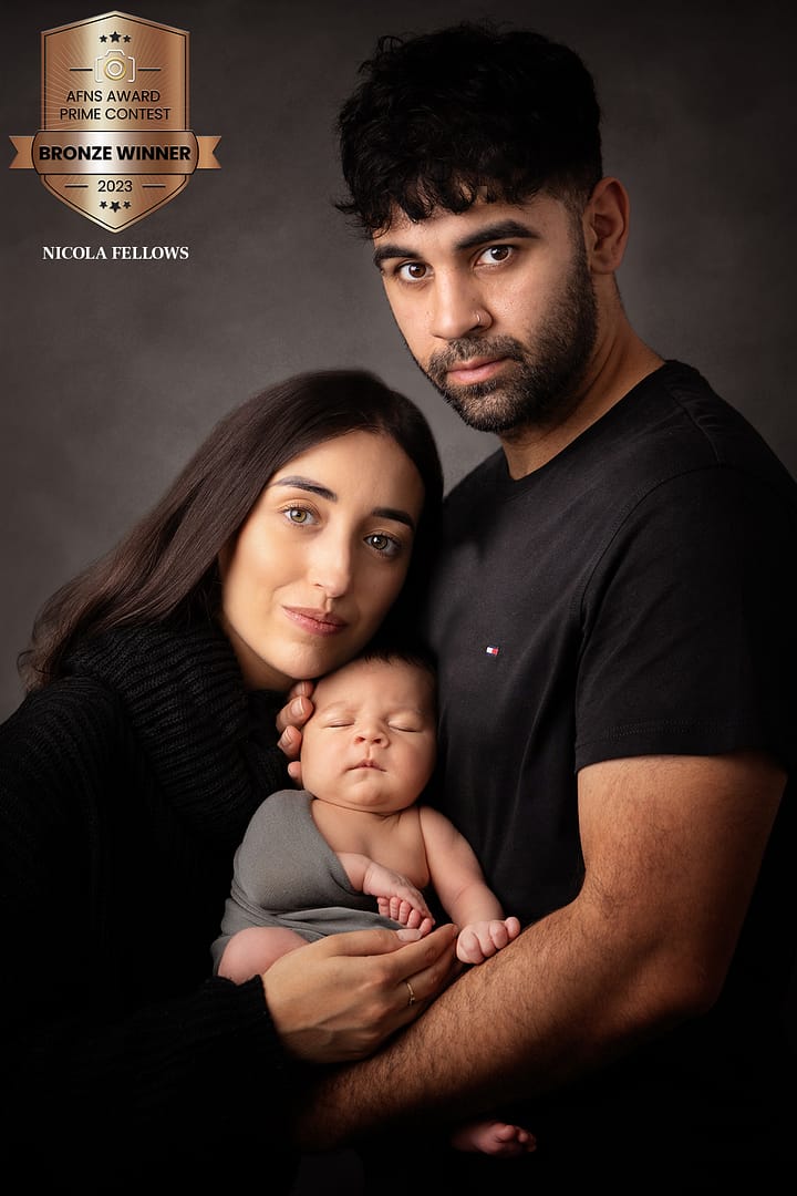 newborn baby boy wrapped in a grey wrap is being held by his Mom and Dad, who are both dressed in black. Taken by newborn photographer Nicki Byrne Photography, newborn photographer birmingham
