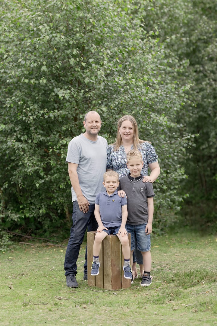Situated in Sutton Park, Outdoor family photographer Nicki Byrne Photography has captured this family of four - Mom, Dad and two young sons - standing/sitting together smiling at the camera. Family photographer Sutton Coldfield birmingham